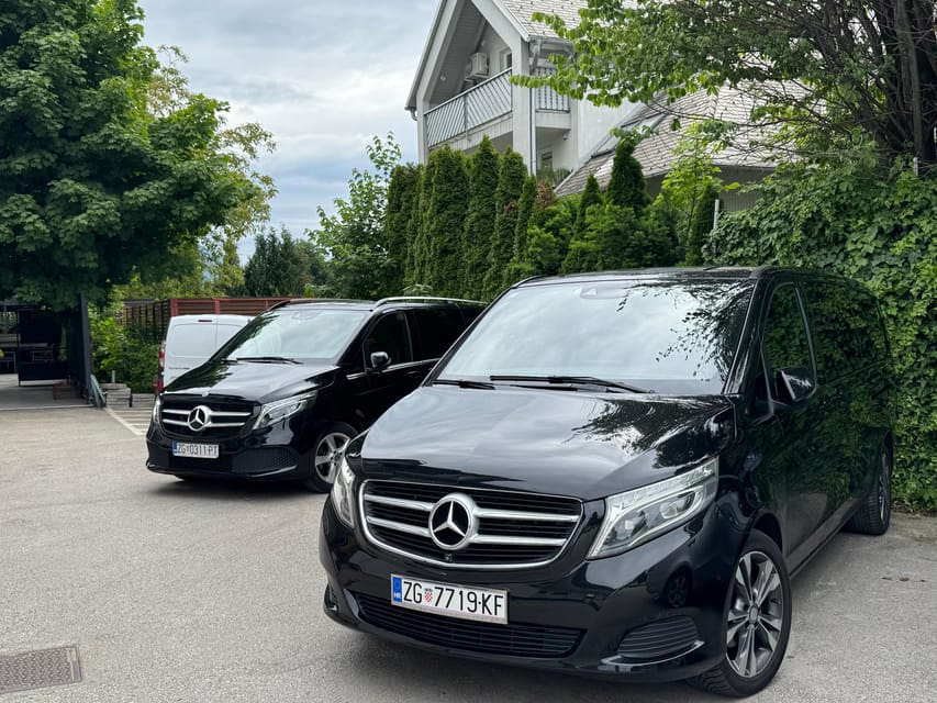 Two black Mercedes minibuses parked in a car park beside green trees and a house