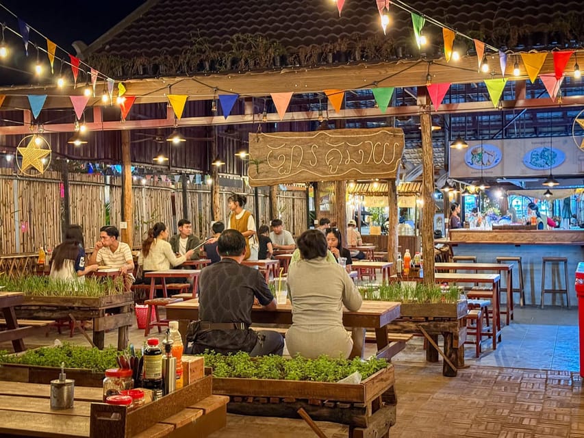 An evening eating place under a roof strung with bunting and light bulbs, guests at wooden tables with grasses planted in crates between them, a hand-painted Khmer sign above