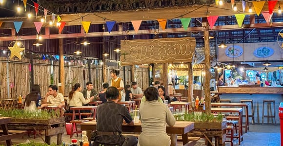 An evening eating place under a roof strung with bunting and light bulbs, guests at wooden tables with grasses planted in crates between them, a hand-painted Khmer sign above