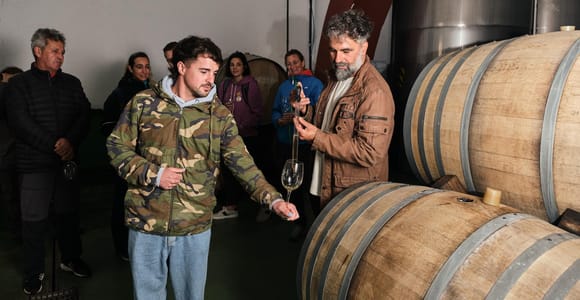 A winery guide draws wine from a wooden barrel in the cellar while a group of visitors watches