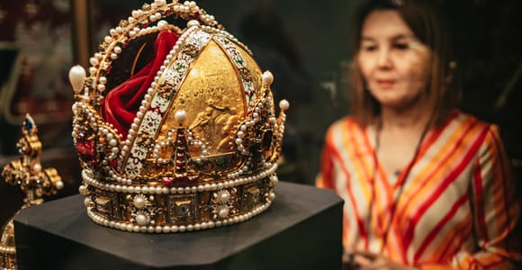 Gold crown set with pearls and gemstones in a display case, a visitor out of focus behind it