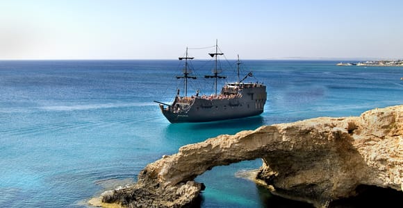A dark wooden pirate ship with three masts sailing past a natural rock arch over turquoise water