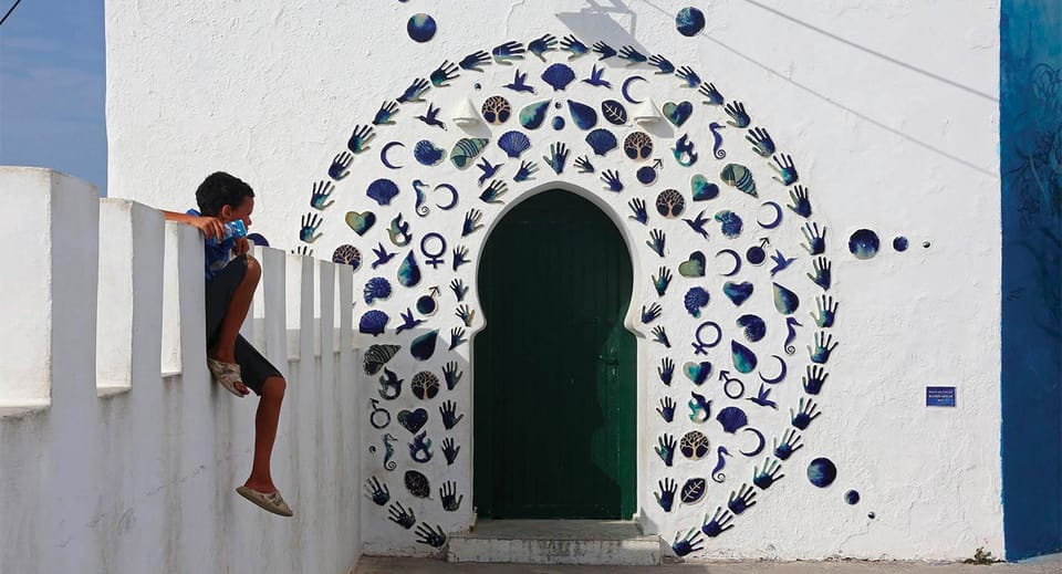 A white wall with a green arched door framed by blue ceramic shells, hands and crescents, a boy sitting on the parapet beside it
