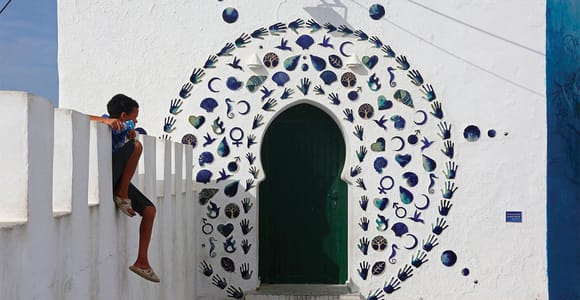 A white wall with a green arched door framed by blue ceramic shells, hands and crescents, a boy sitting on the parapet beside it