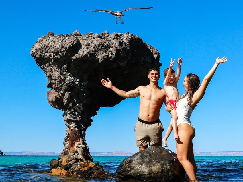 A family posing on a boulder beside the mushroom-shaped rock in shallow turquoise water, a gull overhead and pale desert hills across the bay