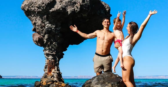 A family posing on a boulder beside the mushroom-shaped rock in shallow turquoise water, a gull overhead and pale desert hills across the bay