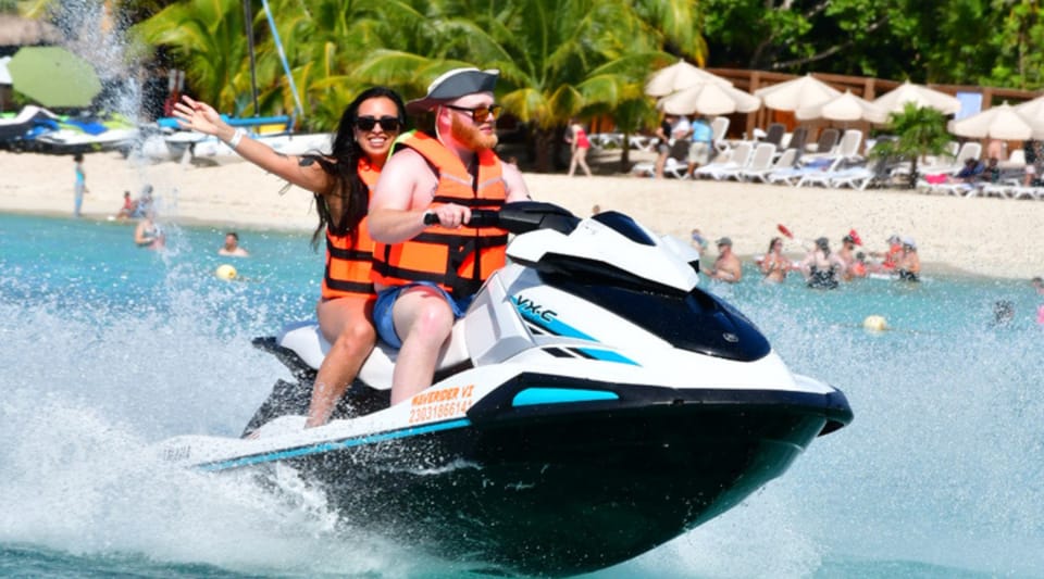 Two people in orange life jackets riding a jet ski through the spray, with sun loungers, parasols and palms on the beach behind