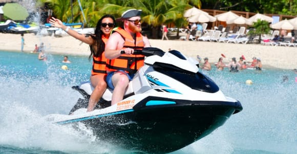 Two people in orange life jackets riding a jet ski through the spray, with sun loungers, parasols and palms on the beach behind