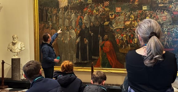 A guide points at a large gilded-frame painting while visitors, including children, look on beside a stone bust