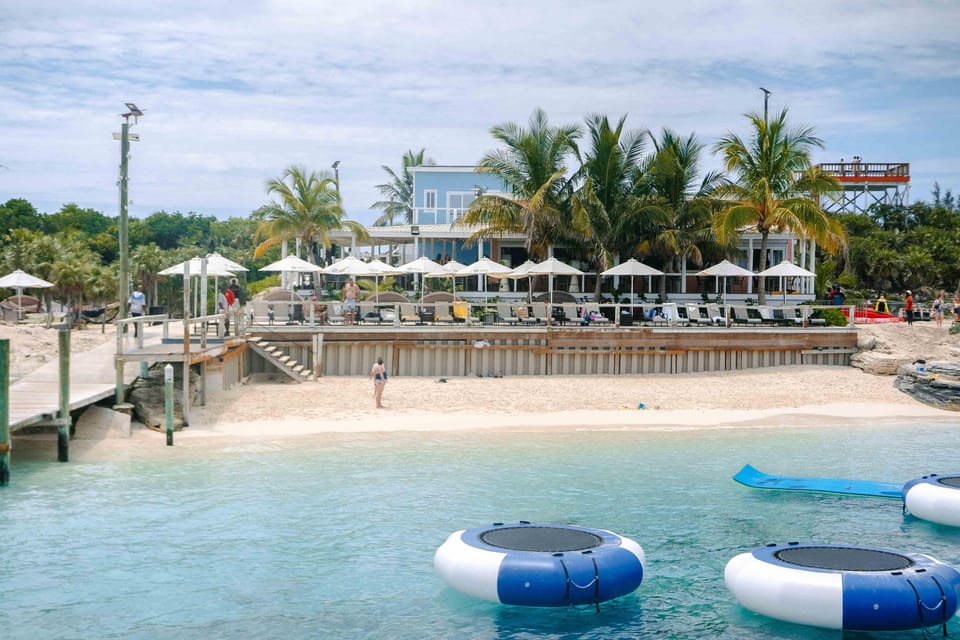 A Rose Island beach club seen from the water: palm trees and thatched umbrellas above a low sea wall, a wooden jetty on the left, and two blue-and-white inflatable water trampolines floating in the shallows