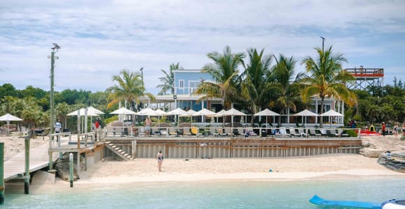 A Rose Island beach club seen from the water: palm trees and thatched umbrellas above a low sea wall, a wooden jetty on the left, and two blue-and-white inflatable water trampolines floating in the shallows