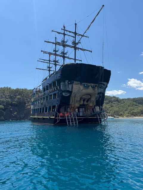 Large pirate-style wooden boat anchored in a turquoise bay below pine-covered hills