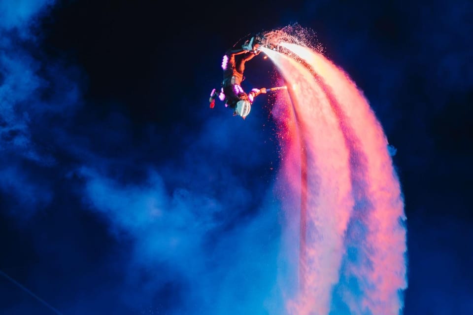 A flyboard performer lit pink against dark blue smoke above the water, twin jets of spray arcing below