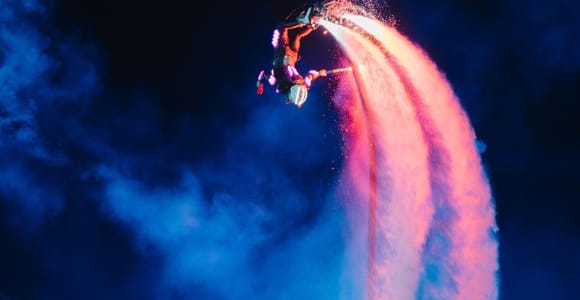 A flyboard performer lit pink against dark blue smoke above the water, twin jets of spray arcing below