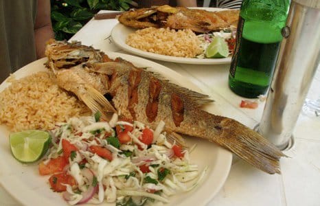 Two plates of whole fried fish with rice, a chopped tomato and onion salad and lime wedges on a white tablecloth, a green beer bottle standing beside them