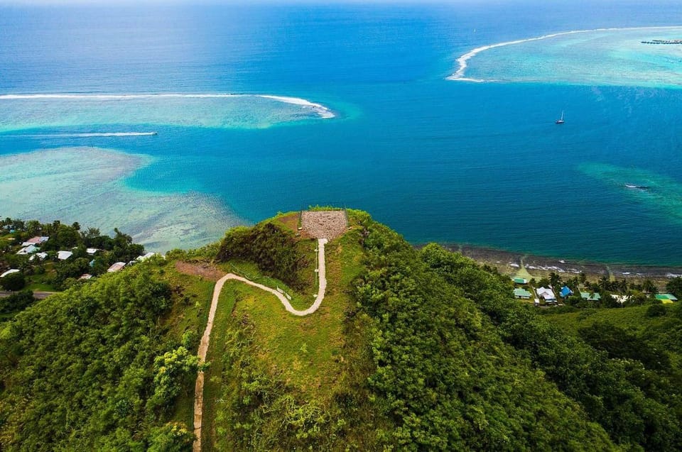 Aerial view of a green headland with a small lookout platform and a pale track leading to it, a turquoise lagoon, a white line of reef break and deep blue ocean beyond