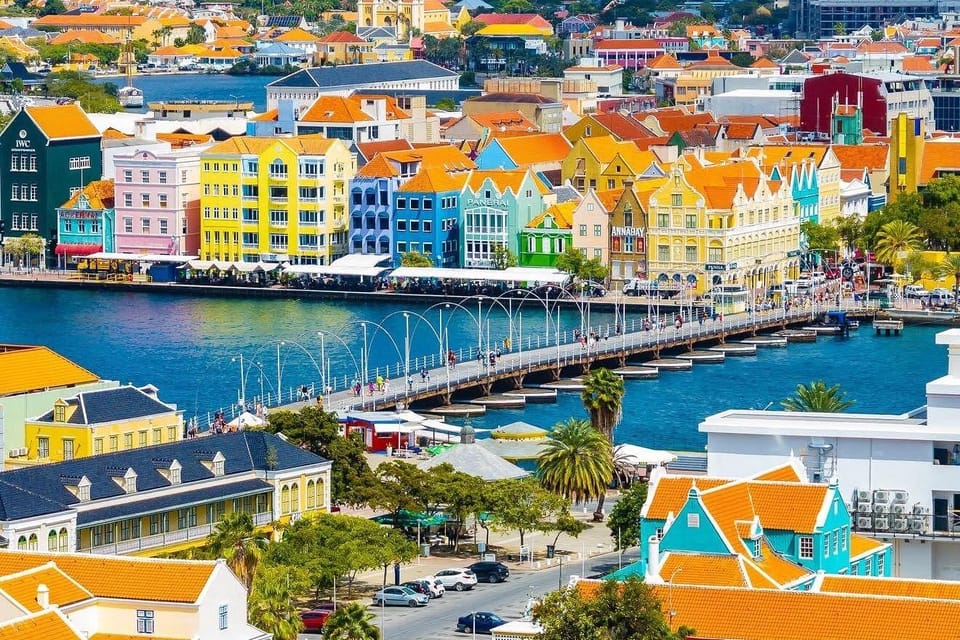 Willemstad seen from above: a row of pastel gabled merchant houses with orange tiled roofs along the waterfront, a long pontoon bridge curving across the bay towards them, and more tiled roofs among palms in the foreground