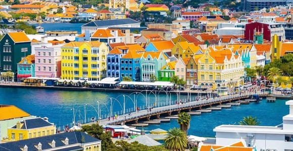 Willemstad seen from above: a row of pastel gabled merchant houses with orange tiled roofs along the waterfront, a long pontoon bridge curving across the bay towards them, and more tiled roofs among palms in the foreground
