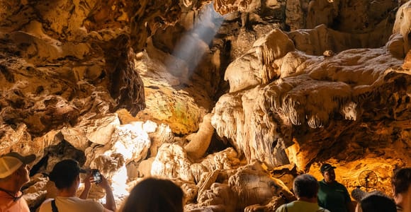 Inside a limestone cave: visitors in a group looking up at ochre stalactites and flowstone, with a shaft of daylight falling through an opening in the roof