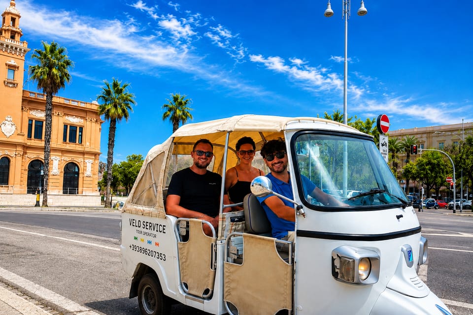 A white three-wheeled Ape tuk-tuk with a driver and two passengers on a sunny street lined with palm trees