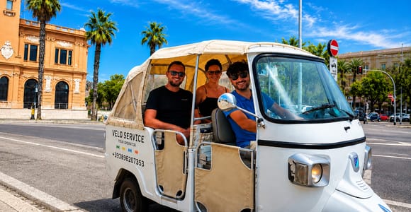 A white three-wheeled Ape tuk-tuk with a driver and two passengers on a sunny street lined with palm trees