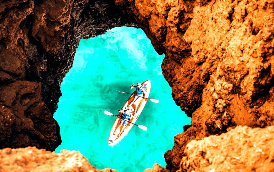 Two people in a double kayak on turquoise water, seen from above through a rock opening in an orange cliff