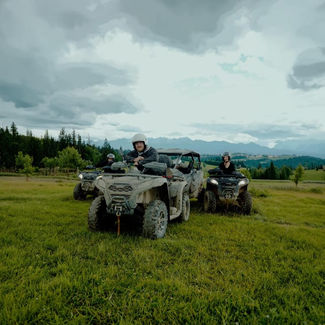Muddy quad bikes and a buggy parked on a meadow, riders in helmets, Tatra ridge behind