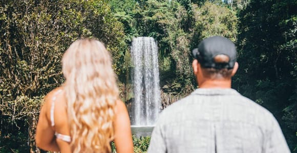 Two people seen from behind, a woman with long fair hair and a man in a dark cap, watching a single tall waterfall drop into a dark pool surrounded by dense green forest