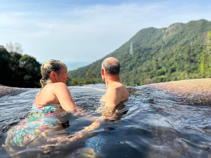 Zwei Menschen sitzen schulterhoch in einem natürlichen Felsbecken auf einer nackten Steinplatte und blicken über bewaldete Bergkämme zum Meer, im Hintergrund ist knapp eine Seilbahnstrecke am Berghang zu erkennen
