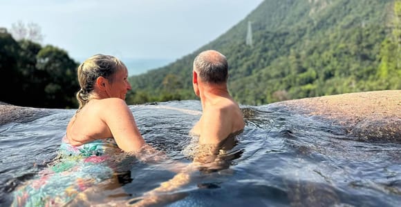 Two people sitting shoulder-deep in a natural rock pool on a bare slab, looking out over forested ridges towards the sea, a cable car line just visible on the mountainside