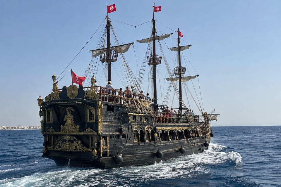 A dark wooden sailing ship with three masts and red flags, passengers along the deck, on open blue water