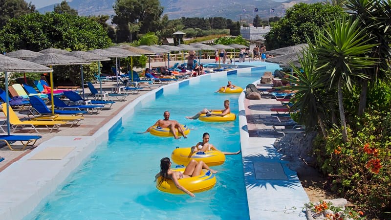 Swimmers on yellow rubber rings drifting along a wide blue lazy river lined with sun loungers and thatched parasols