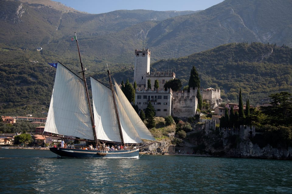 A historic two-masted sailing boat with white sails passing the grey stone castle of Malcesine, with wooded mountains rising behind it