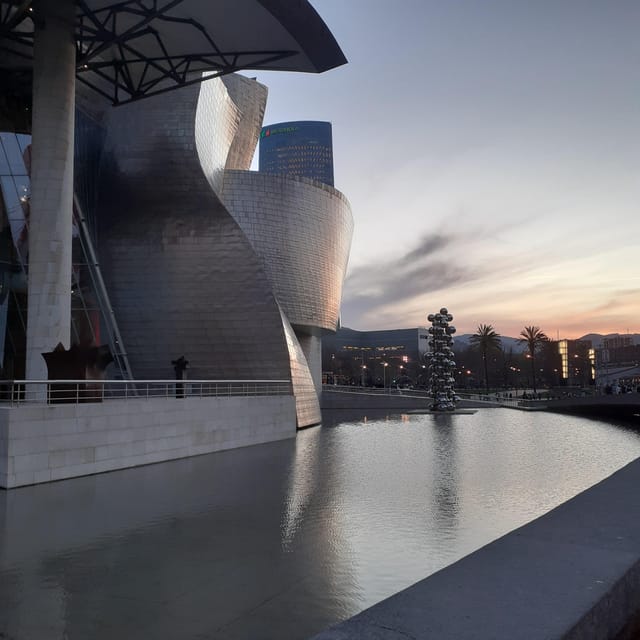 The curved titanium walls of the Guggenheim Museum beside a still pool at dusk, with a sculpture of silver spheres beyond