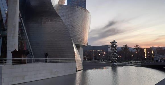 The curved titanium walls of the Guggenheim Museum beside a still pool at dusk, with a sculpture of silver spheres beyond
