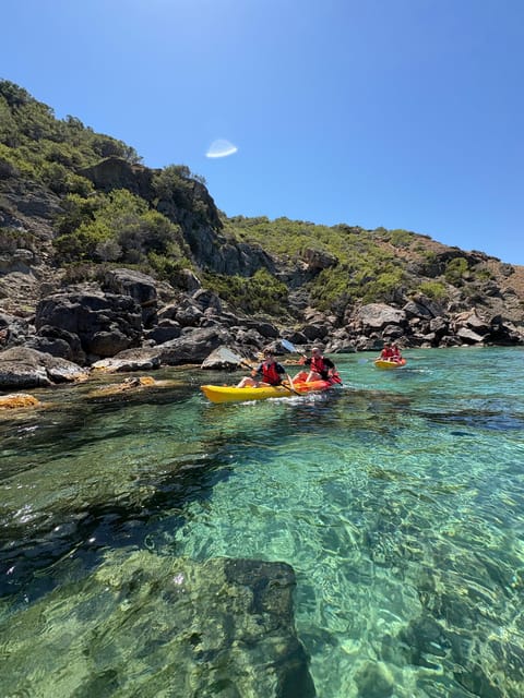 Two people in life jackets paddling a yellow kayak on clear green water below a scrubby rocky shore