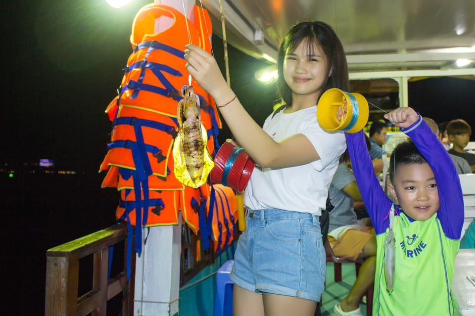 A young woman on a lit boat deck at night holding up a squid on a line, a child beside her holding a second one, orange life jackets hanging behind
