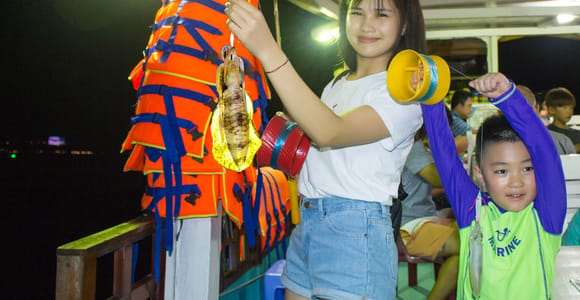 A young woman on a lit boat deck at night holding up a squid on a line, a child beside her holding a second one, orange life jackets hanging behind