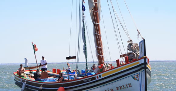 A traditional Portuguese wooden boat named SEJAS FELIZ sailing on the open Tagus, several guests on board and the Portuguese flag at the stern