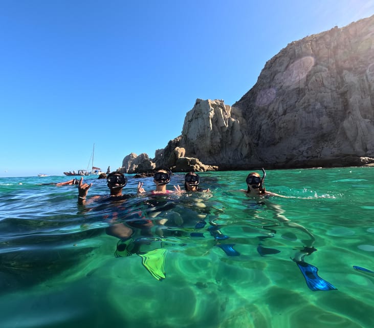 Four snorkellers in masks at the surface of clear green water below the rock cliffs of Land's End, a boat anchored behind them