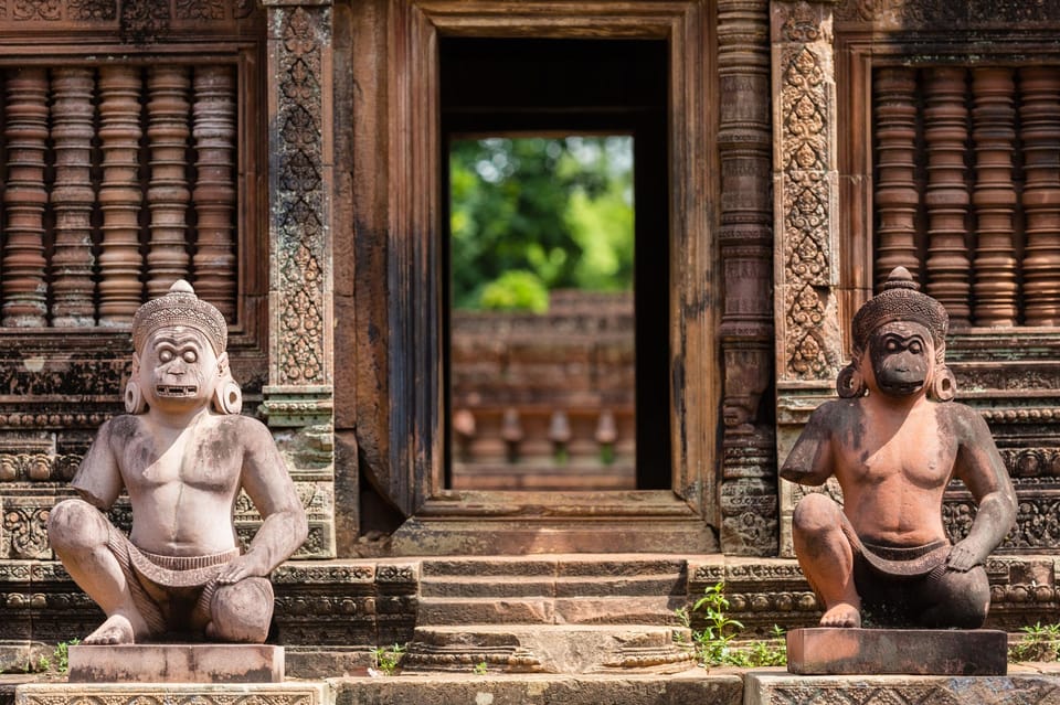 Two squatting guardian figures on plinths flanking a temple doorway, the surrounding pink sandstone covered in carved foliage and baluster windows