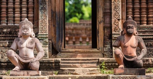 Two squatting guardian figures on plinths flanking a temple doorway, the surrounding pink sandstone covered in carved foliage and baluster windows