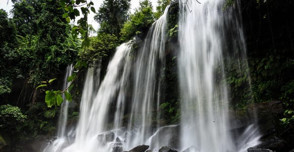 A wide waterfall dropping into a brown pool ringed by jungle, a few people standing on the wet rocks at the bottom