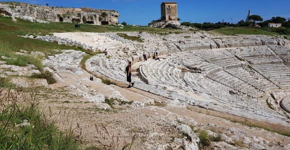 Curved tiers of white stone seating in an ancient Greek theatre on a grassy hillside under a blue sky