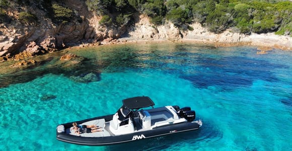A large black and white RIB at anchor in clear turquoise water beside a rocky, wooded shore