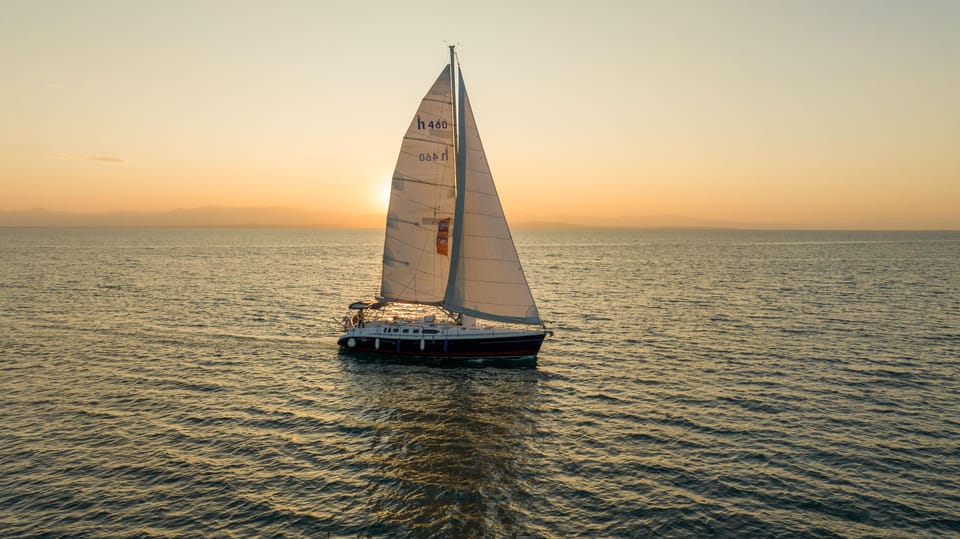 A white sailing yacht on calm water with a low orange sun and a hazy horizon behind it