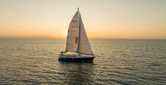 A white sailing yacht on calm water with a low orange sun and a hazy horizon behind it