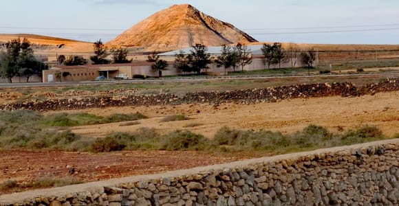 Ein sonnenbeschienener Vulkankegel über flachem Ackerland und niedrigen Gebäuden, im Vordergrund eine Trockenmauer