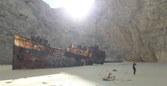 A rusty shipwreck lies on a sandy beach below tall pale cliffs, with a person standing in front