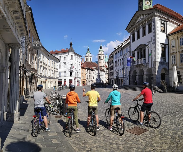 Five cyclists in helmets stand with their bikes on a cobbled square, facing pastel old town houses and church towers under a blue sky